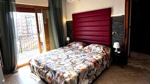 Een slaapkamer met een bed, een raam met gordijnen, een airconditioning, een nachtkastje met een lamp en een deur.