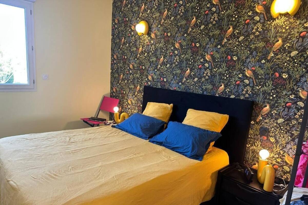 A bed with a yellow and blue bedspread, a dark headboard, and a wall with a bird pattern. There are two bedside lamps and a window on the left.