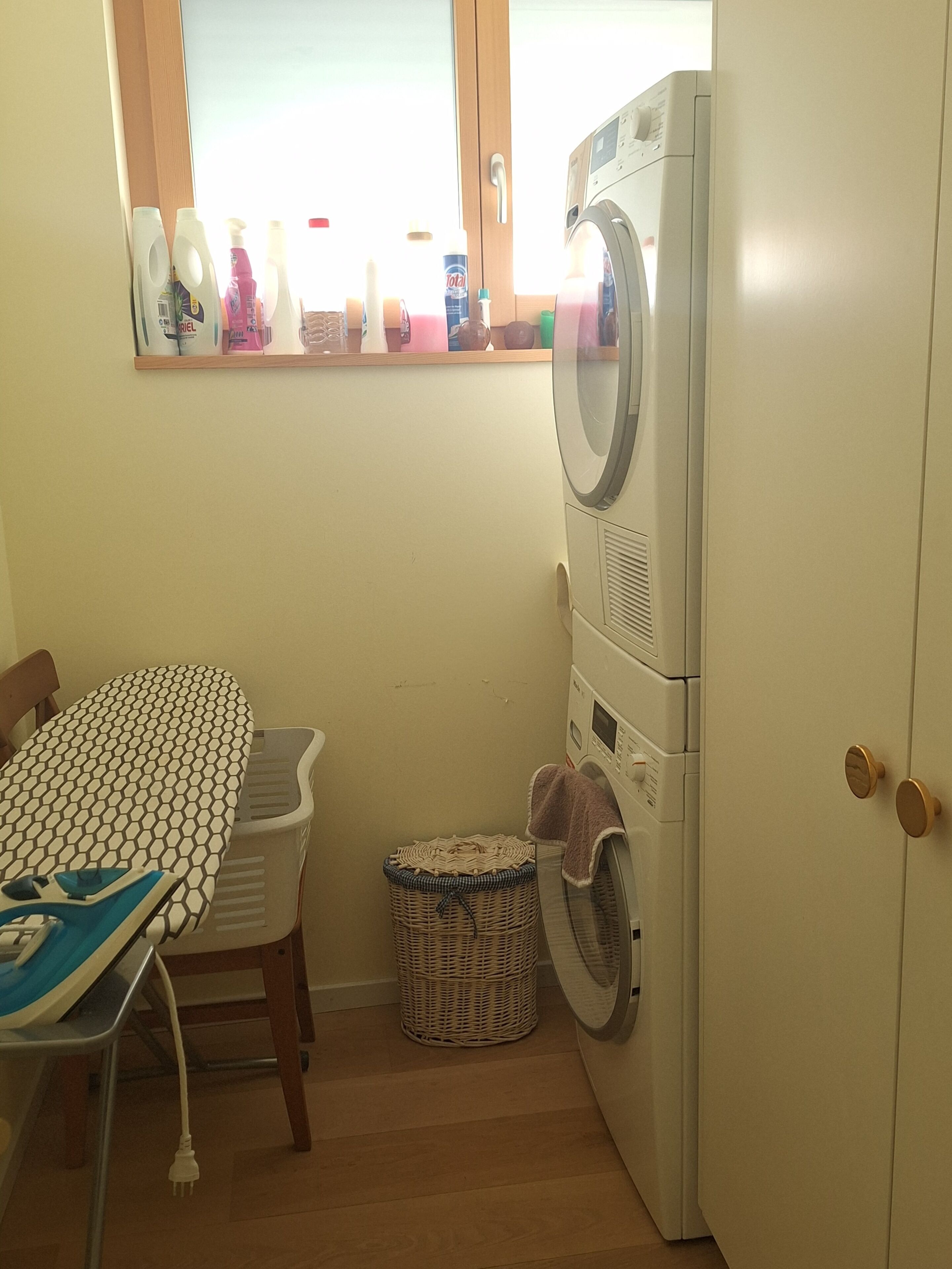 Un espace buanderie comprenant un lave-linge, un étendoir, un panier en osier et une table recouverte d’une nappe à motifs.