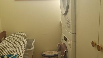 Un espace buanderie comprenant un lave-linge, un étendoir, un panier en osier et une table recouverte d’une nappe à motifs.