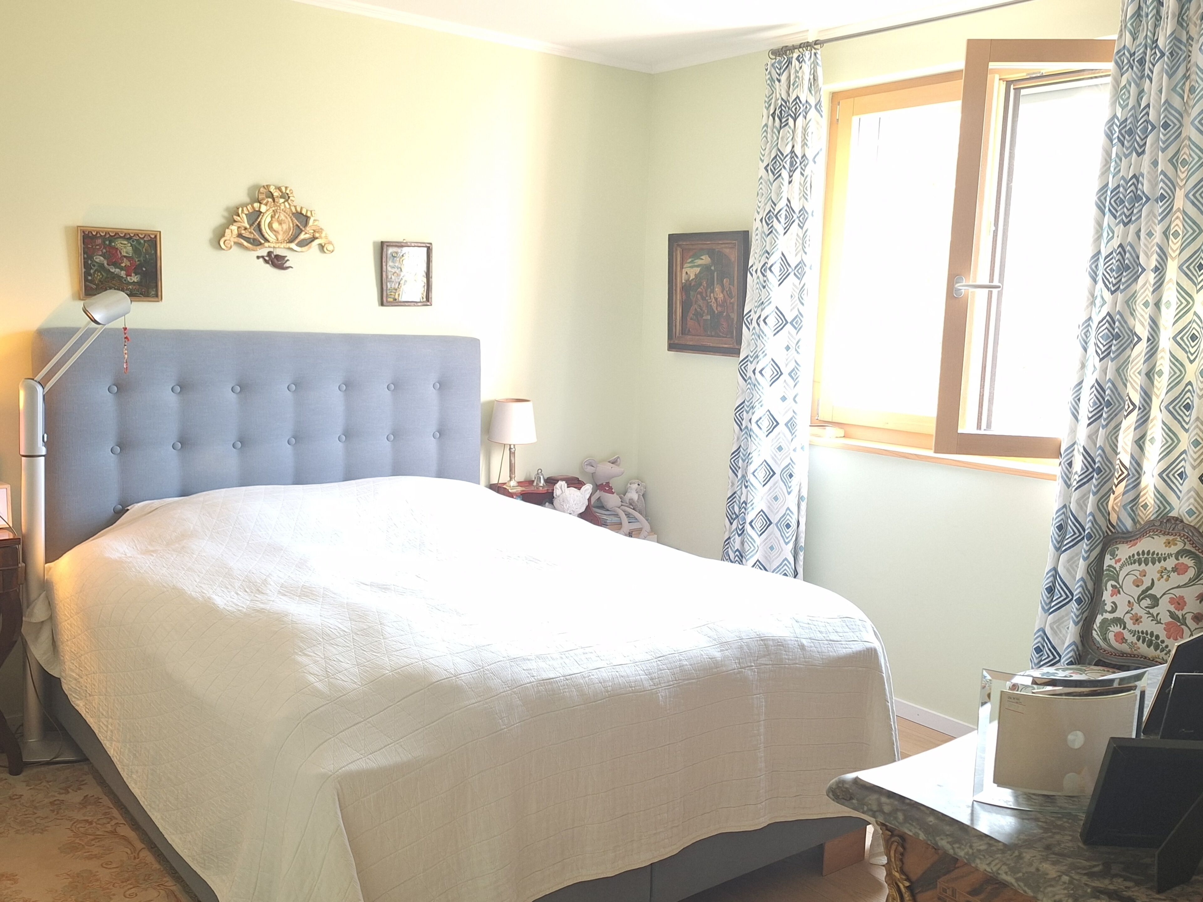 Une chambre à coucher avec un grand lit, une table de chevet, une lampe et une fenêtre avec des rideaux.