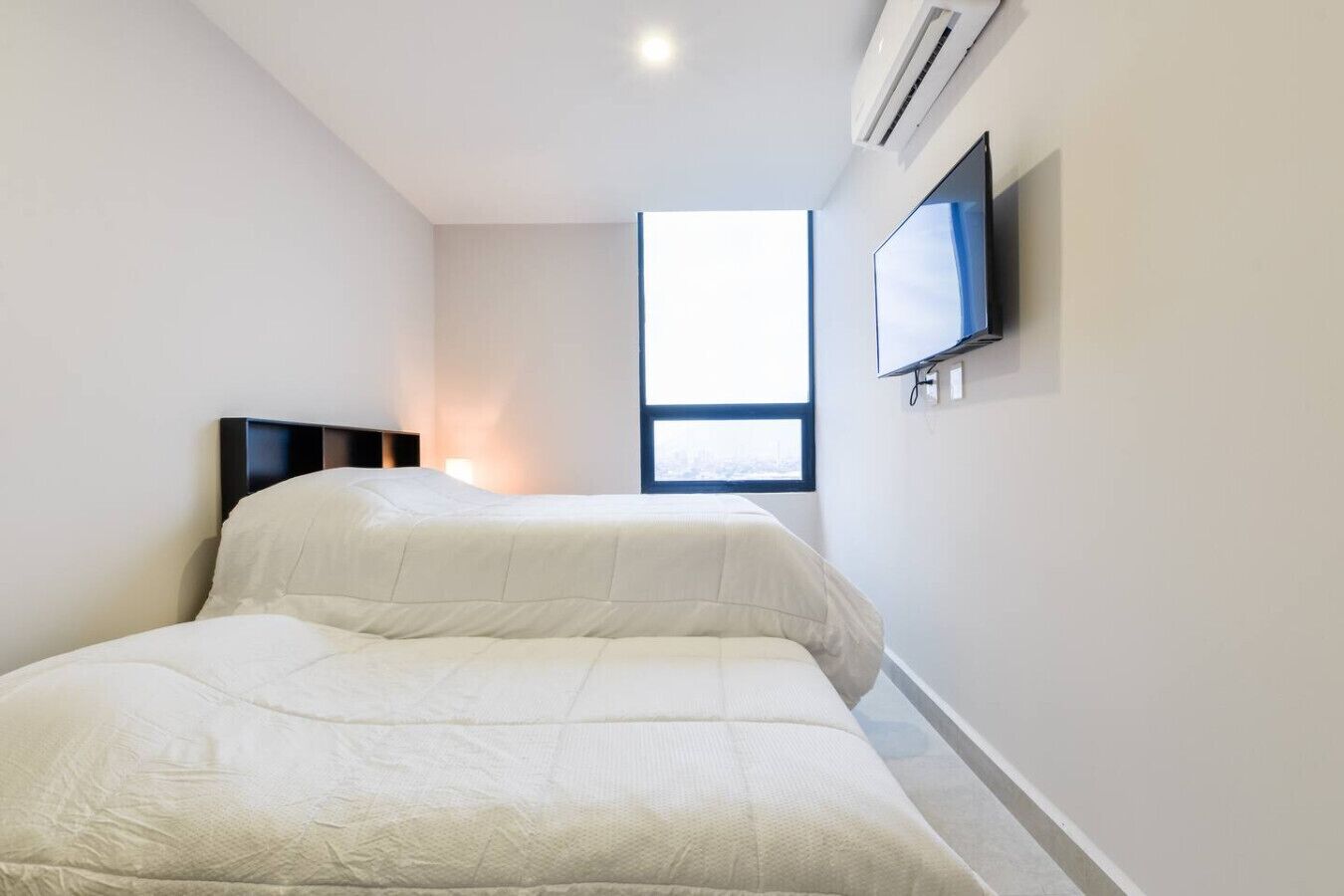 A bedroom with two beds, a TV mounted on the wall, and a window providing natural light.