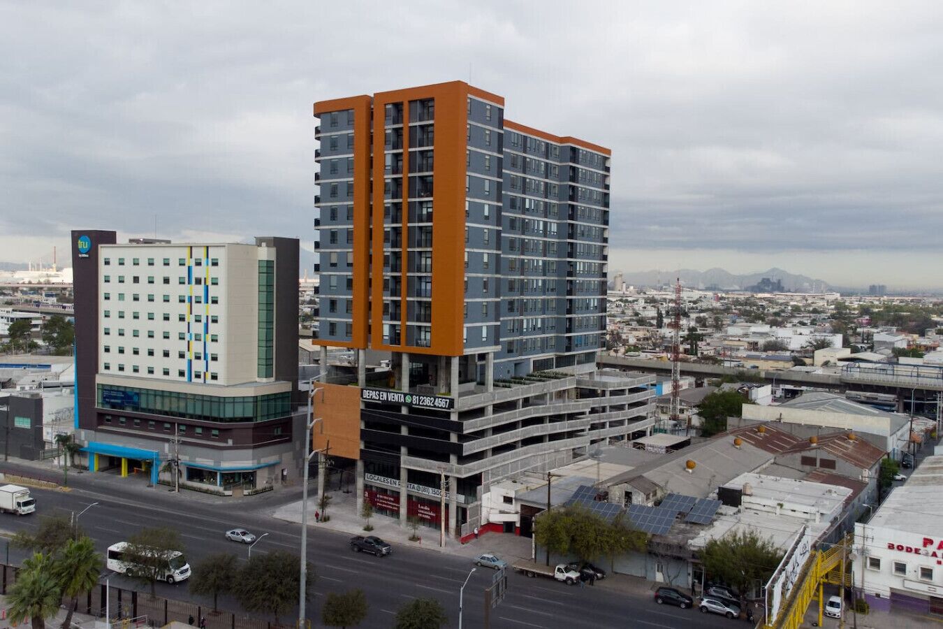 A multi-story hotel with a modern design, featuring a prominent orange and blue facade, a parking garage, and a view of the cityscape in the background.