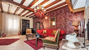 Una sala de estar con un sofá rojo, alfombra estampada, vigas de madera, una araña y un espejo.