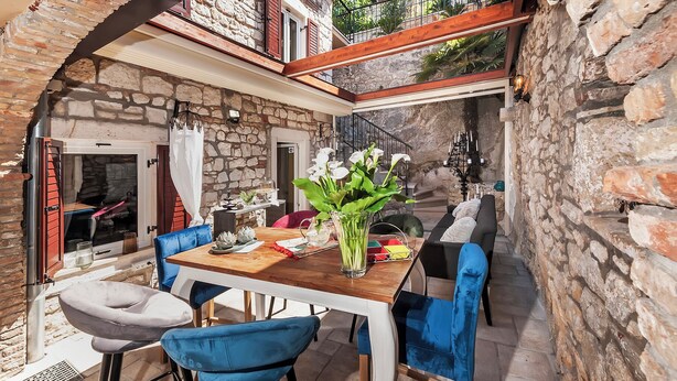 Una habitación con paredes de piedra, una mesa y sillas de madera, un balcón con barandilla y una escalera.
