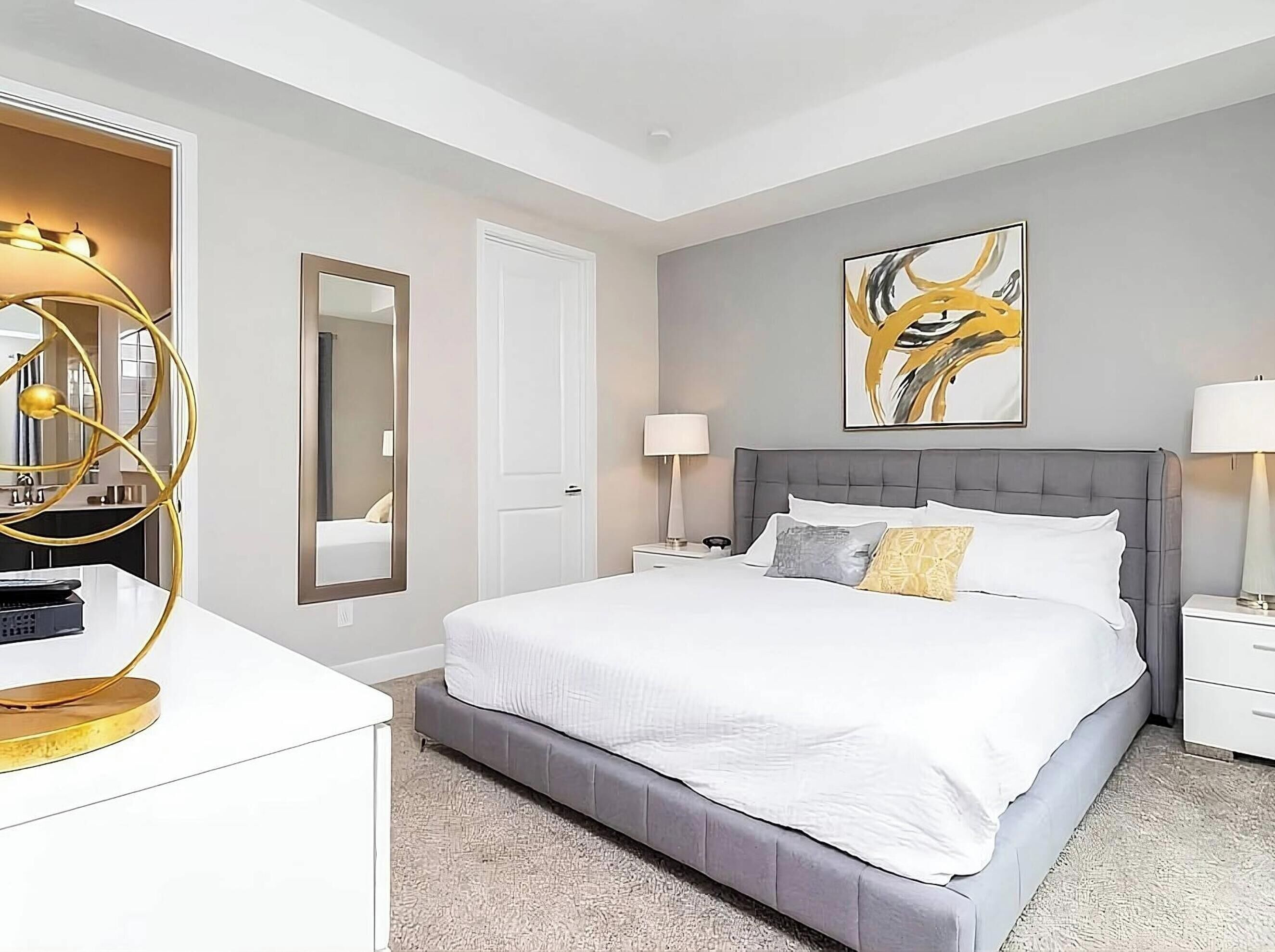 A modern hotel room with a large bed, a bedside table, a nightstand, a mirror, and a wall art piece.