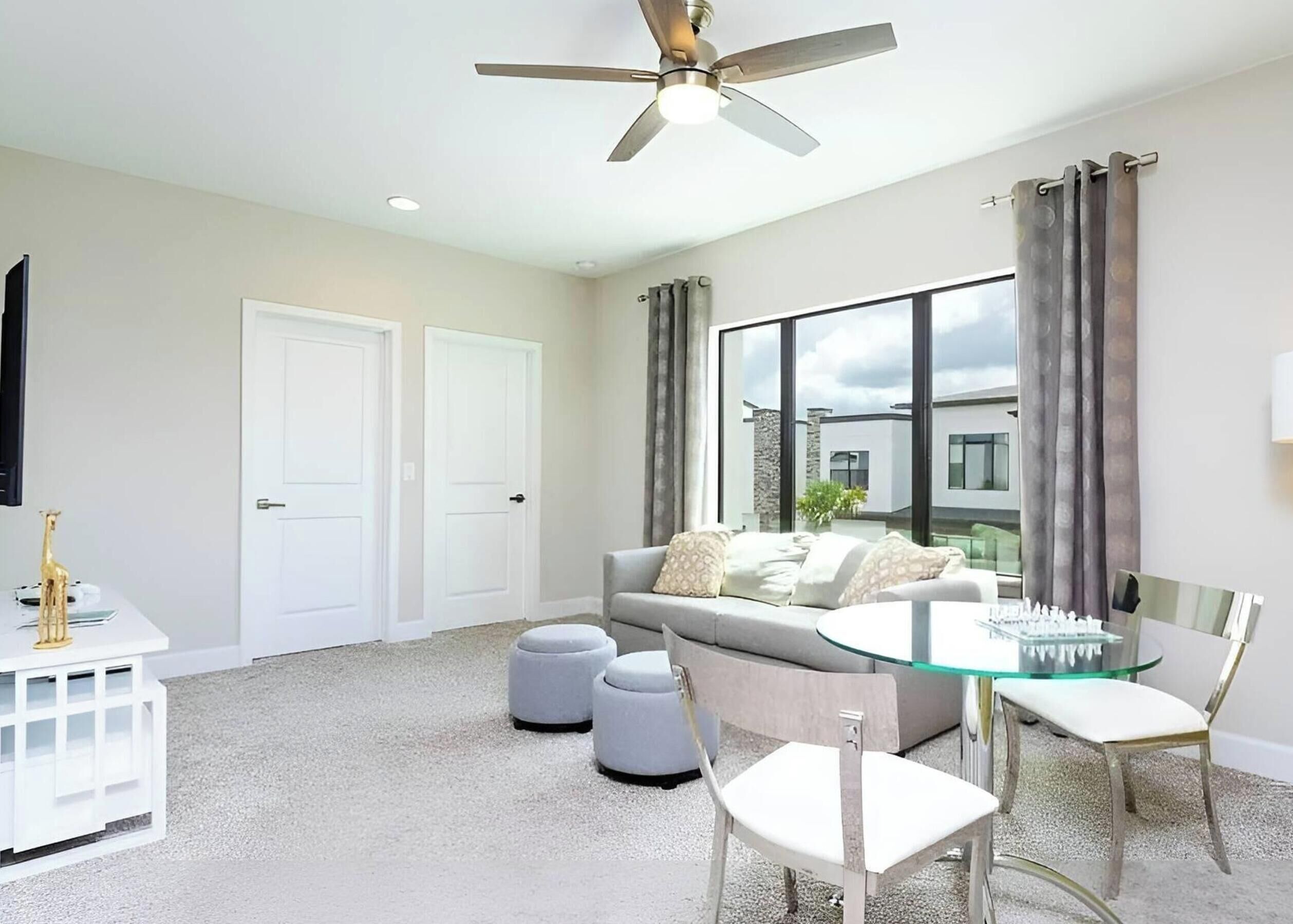 A modern living room with a ceiling fan, a glass table with chairs, a sofa, and a TV mounted on the wall.