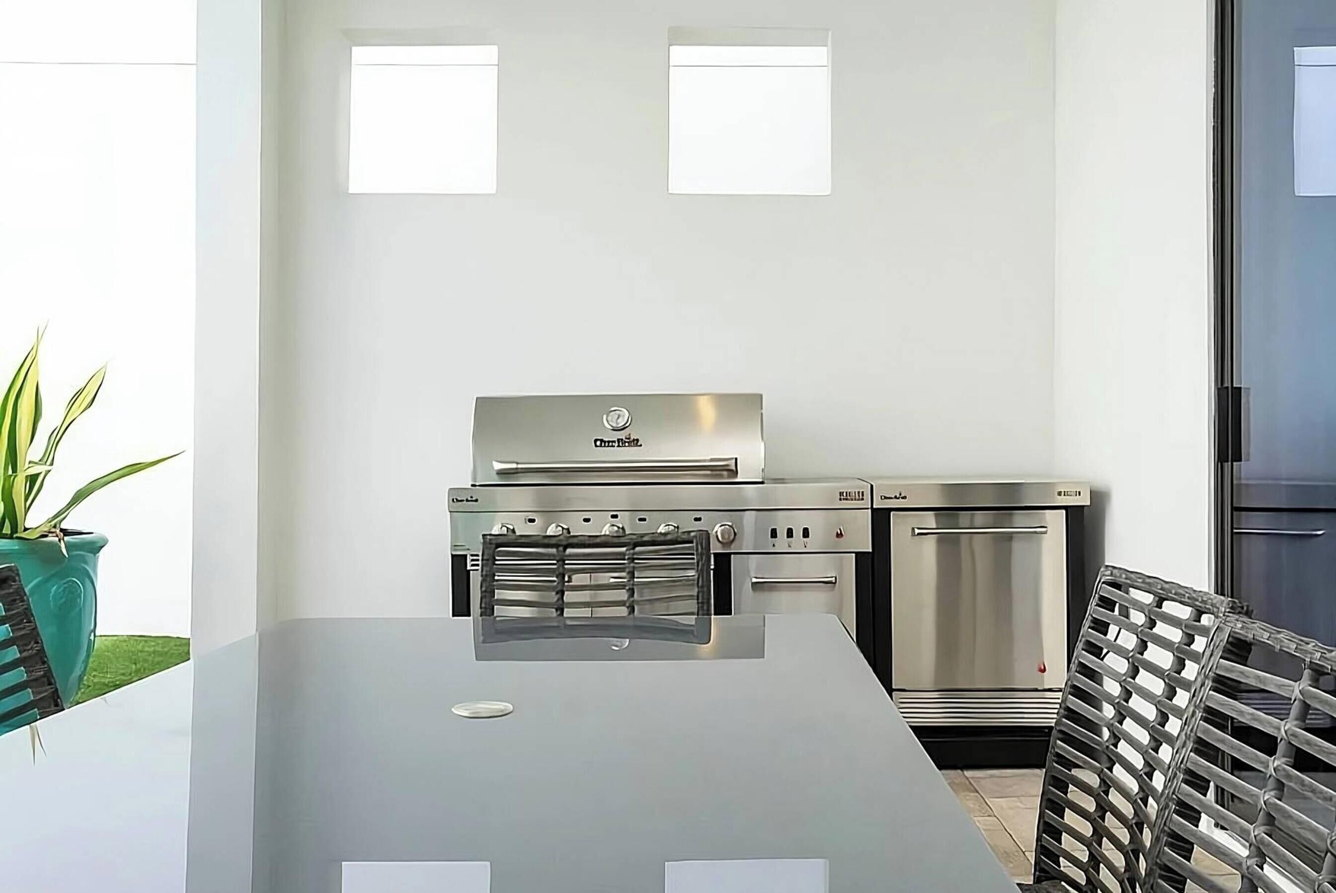 Una cocina con campana extractora de acero inoxidable, horno empotrado y refrigerador. Hay una mesa con una silla metálica y una planta en maceta.