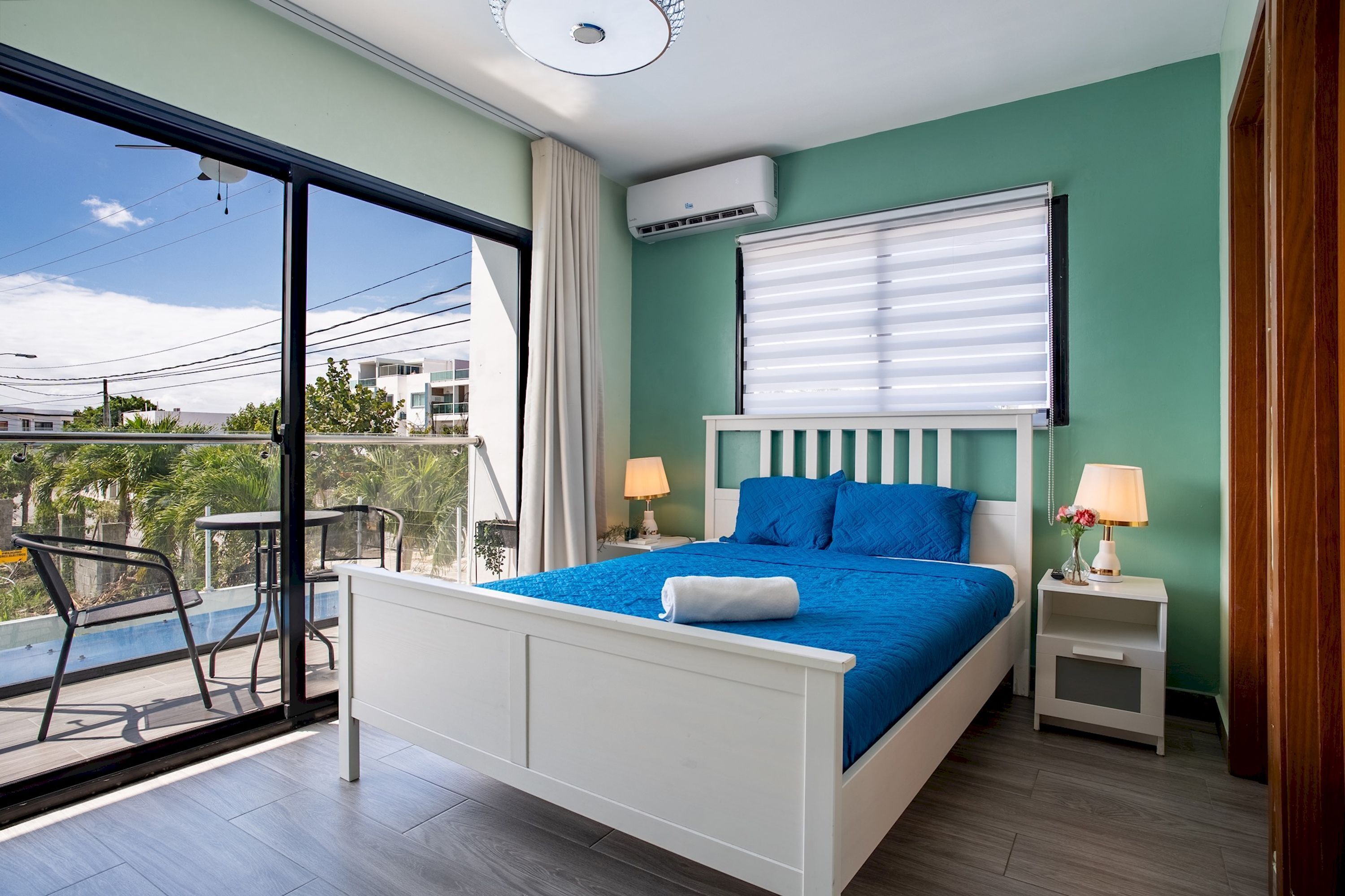 A bedroom with a bed, bedside tables, a window with blinds, an air conditioning unit, and a balcony with a table and chairs.