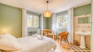 A bedroom with a bed, two chairs, a window, a radiator, a checkered floor, and a bathroom with a sink and mirror. - La Villa Pylataise – Pyla sur mer- Swimming pool (Pyla sur Mer)