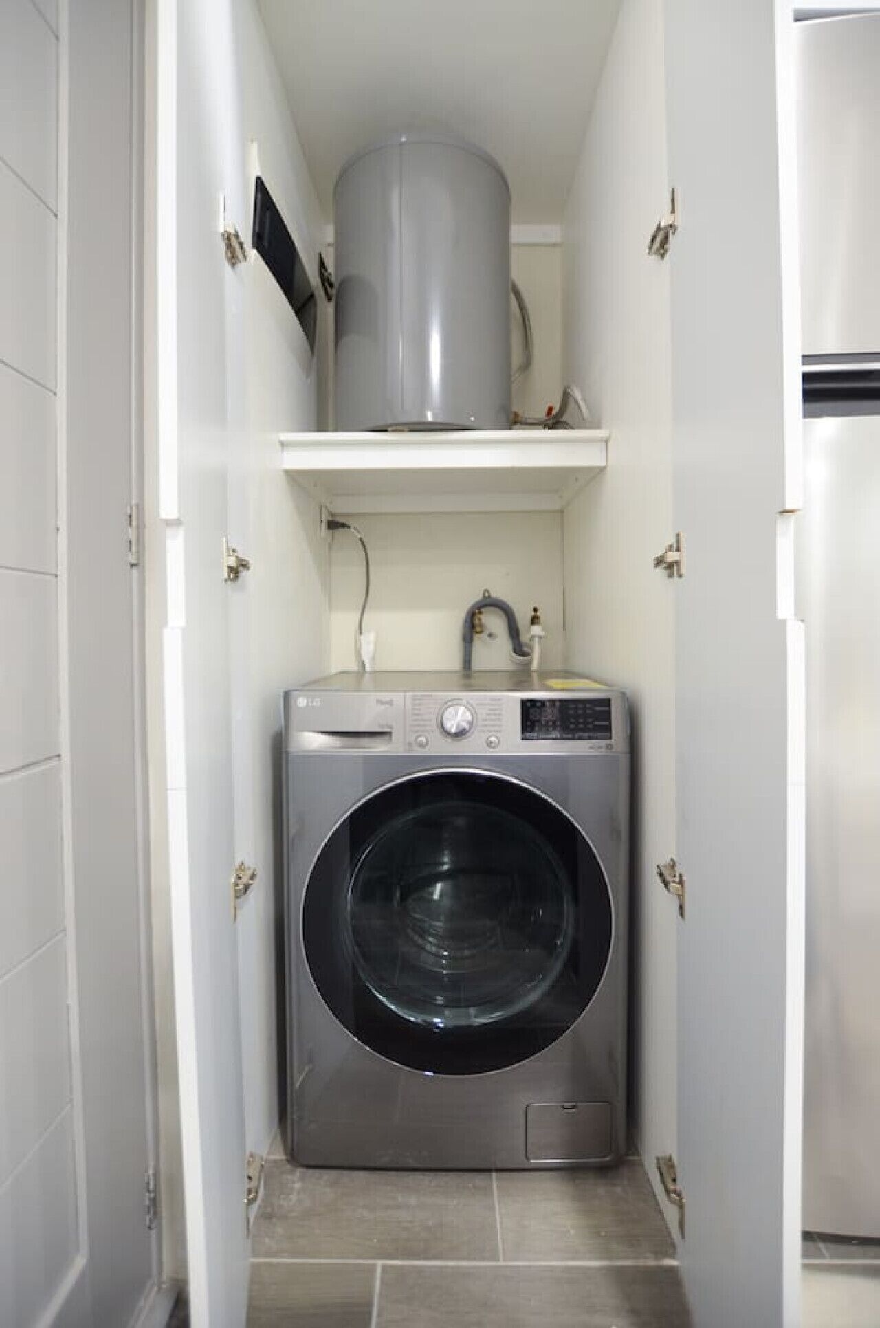 Een kleine wasruimte met een voorlader wasmachine, een klein plankje erboven en een boiler aan de muur.