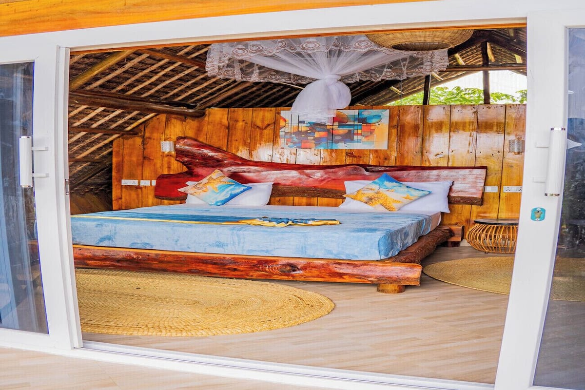 Un dormitorio con cama, almohadas, cabecera de madera, estera tejida y ventilador de techo.