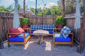 Un espacio de estar al aire libre con madera, con cojines azules y almohadas rojas, rodeado por una valla de bambú y plantas en macetas.