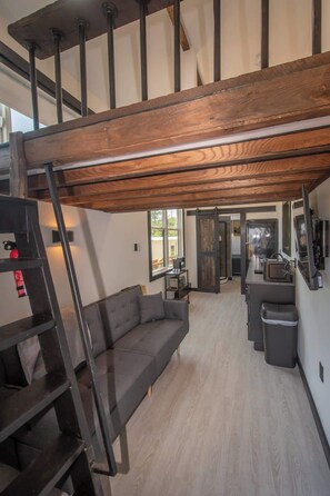 Een loft-achtige woonruimte met een houten plafond, een grijze bank, een keukenhoek en een trap.
