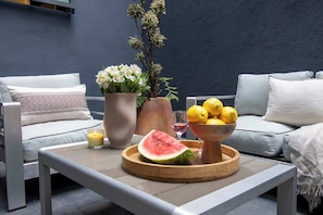 Un espace de détente extérieur moderne, comprenant une table, des coussins, un vase avec des fleurs, un bol de citrons, une tranche de pastèque et une bougie.