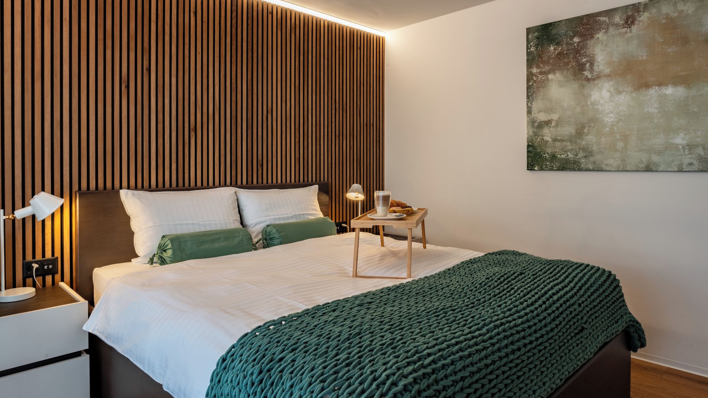 Une chambre moderne avec une tête de lit en lattes de bois, une table de chevet avec une lampe, une autre table de chevet avec un plateau contenant des articles pour le petit-déjeuner, et un plaid vert tricoté sur le lit.