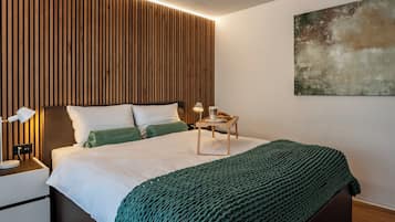 A modern bedroom with a wooden slatted headboard, a bedside table with a lamp, a bedside table with a tray of breakfast items, and a green knitted throw on the bed.