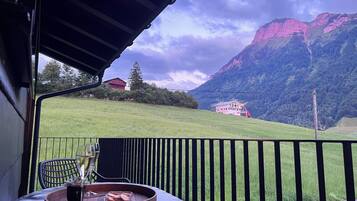 A terrace with a table, chairs, and a glass of water. In the background, there is a mountain and a building.