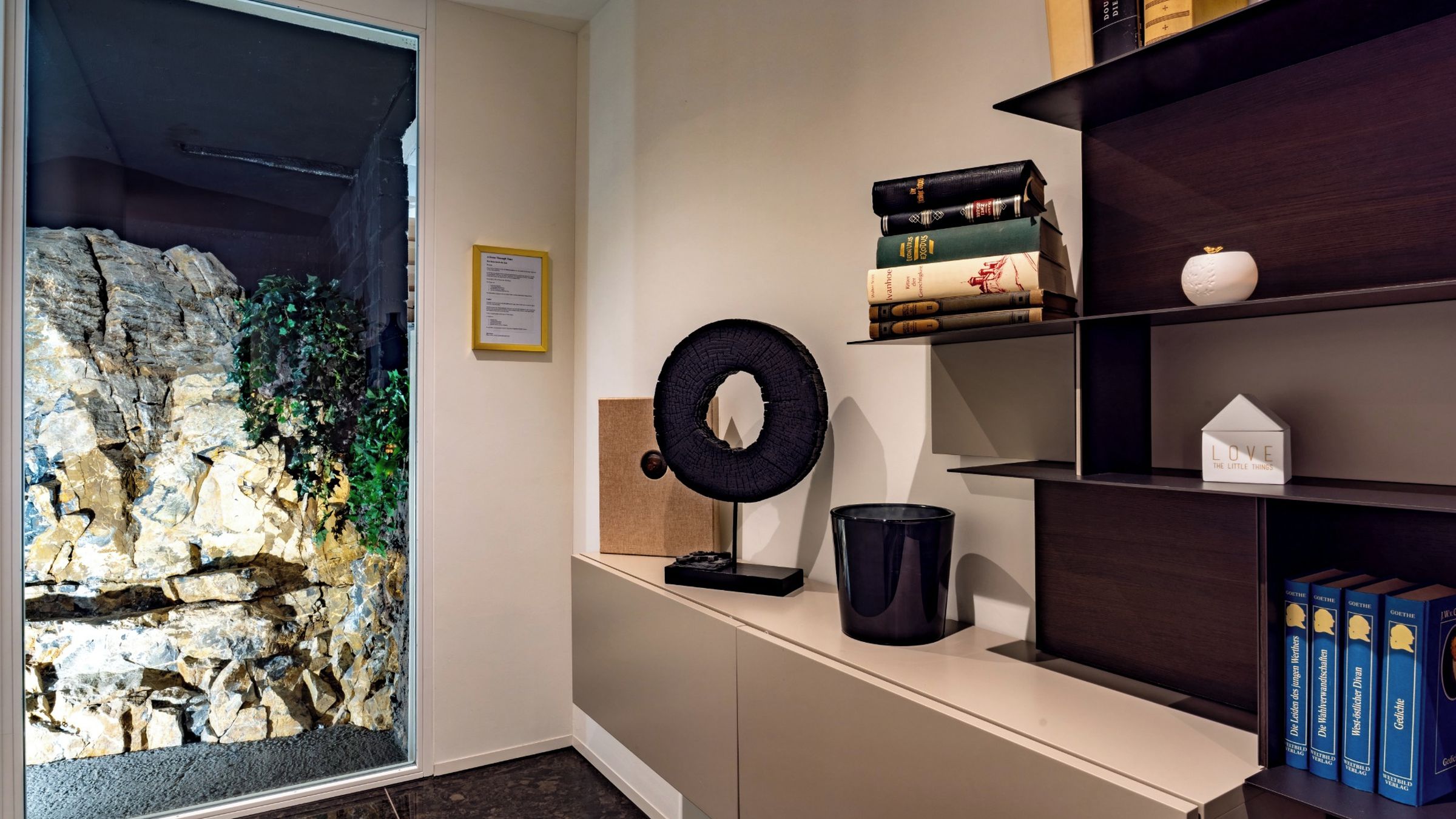 A room with a stone wall feature, a bookshelf with books, a decorative ring, and a small white house-shaped object.