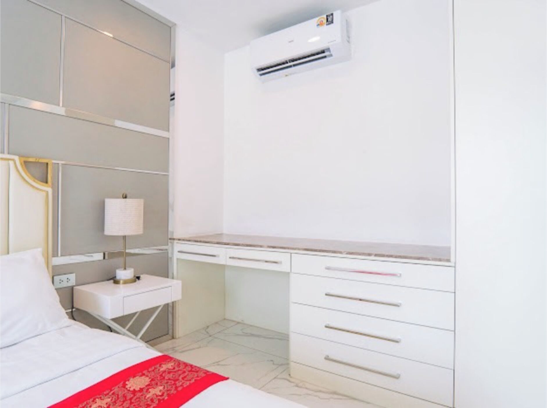 A hotel room with a bed, bedside table, lamp, desk, and air conditioning unit.