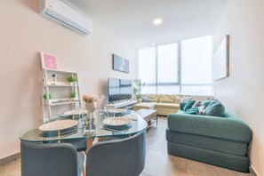 A modern living room with a glass dining table, a sofa, a TV, and a large window.