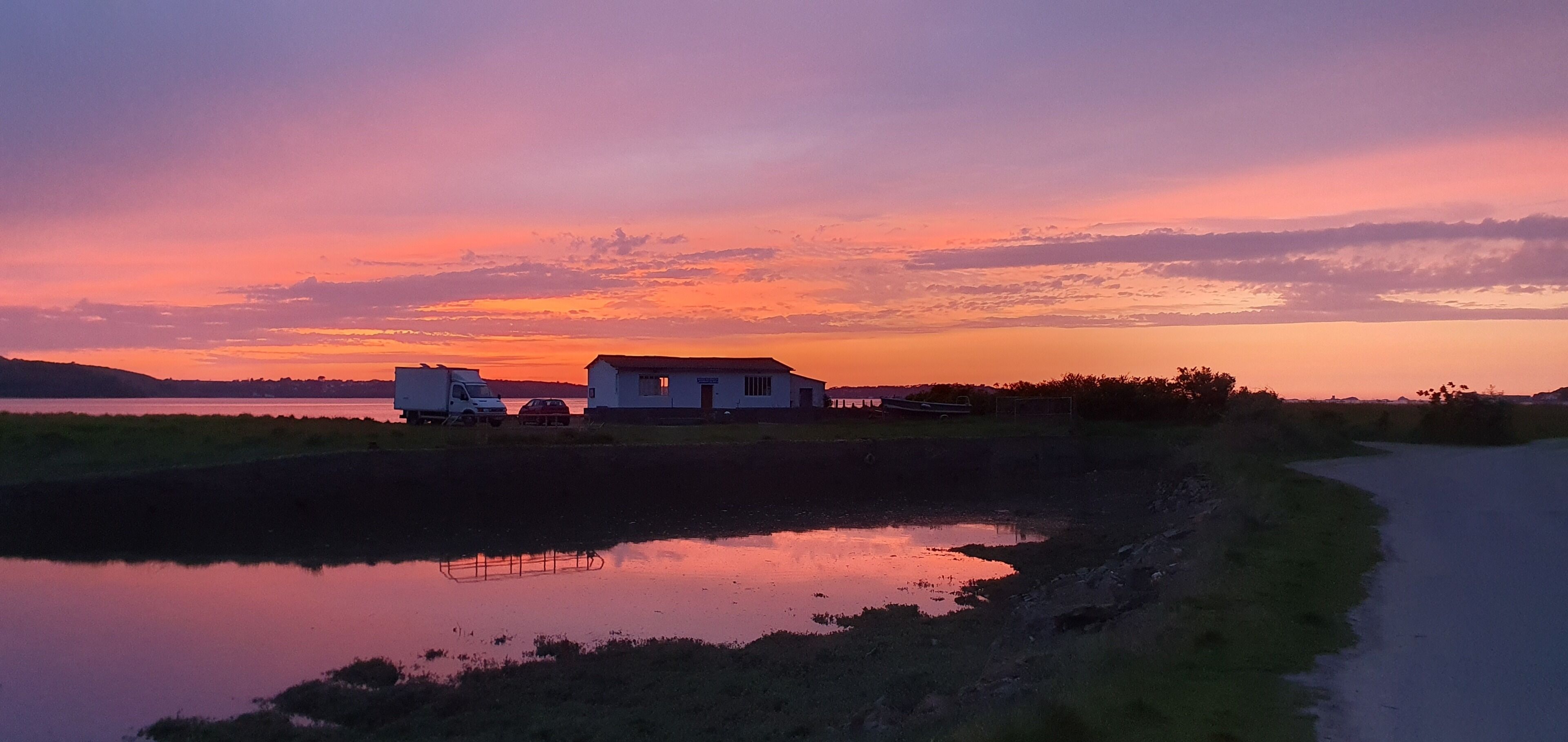 A house with a small pond in front, a road to the right, and a sunset in the background.