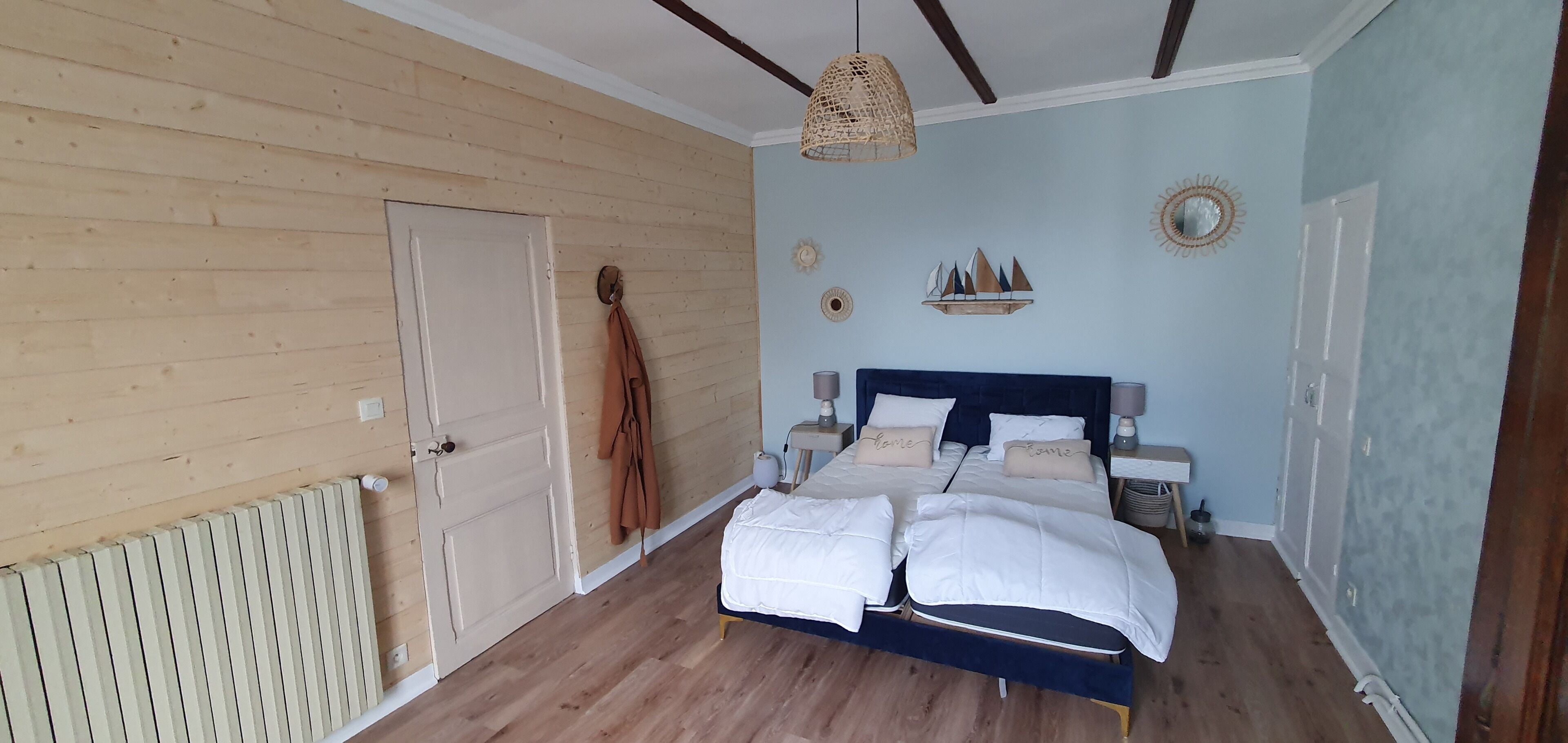Une chambre avec un plancher en bois, un lit avec une literie blanche, un luminaire suspendu et une étagère fixée au mur, ornée d’objets décoratifs.