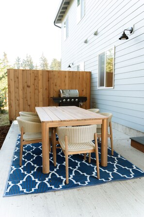 A patio with a wooden table and wicker chairs, a grill, and a blue patterned rug. - The Kat's meow, 3-bedroom duplex in Nehalem (Nehalem)