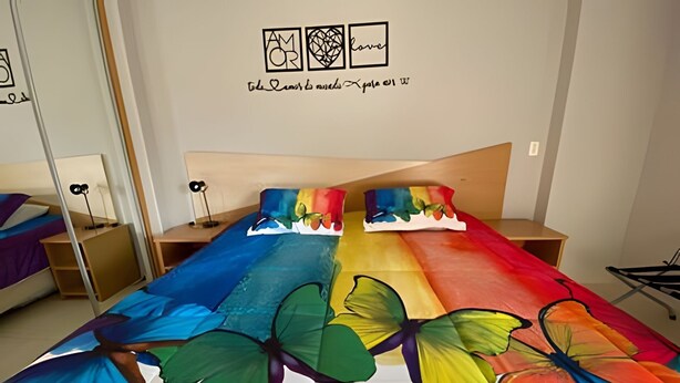 Ein Schlafzimmer mit einem Bett, dessen Bezug bunte Schmetterlingsmotive zeigt, einem Holzkopfende und einer Wand mit gerahmten Kunstwerken.