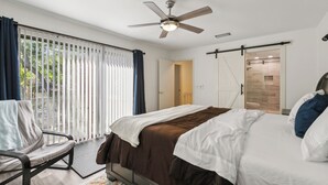A bedroom with a bed, a ceiling fan, a chair, a sliding door to a bathroom, and a view of greenery outside.