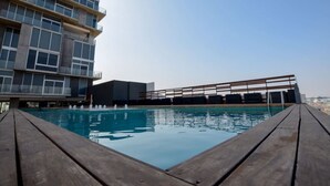 Una piscina sul tetto con un pontile in legno, un edificio moderno con ampie finestre e un cielo sereno.