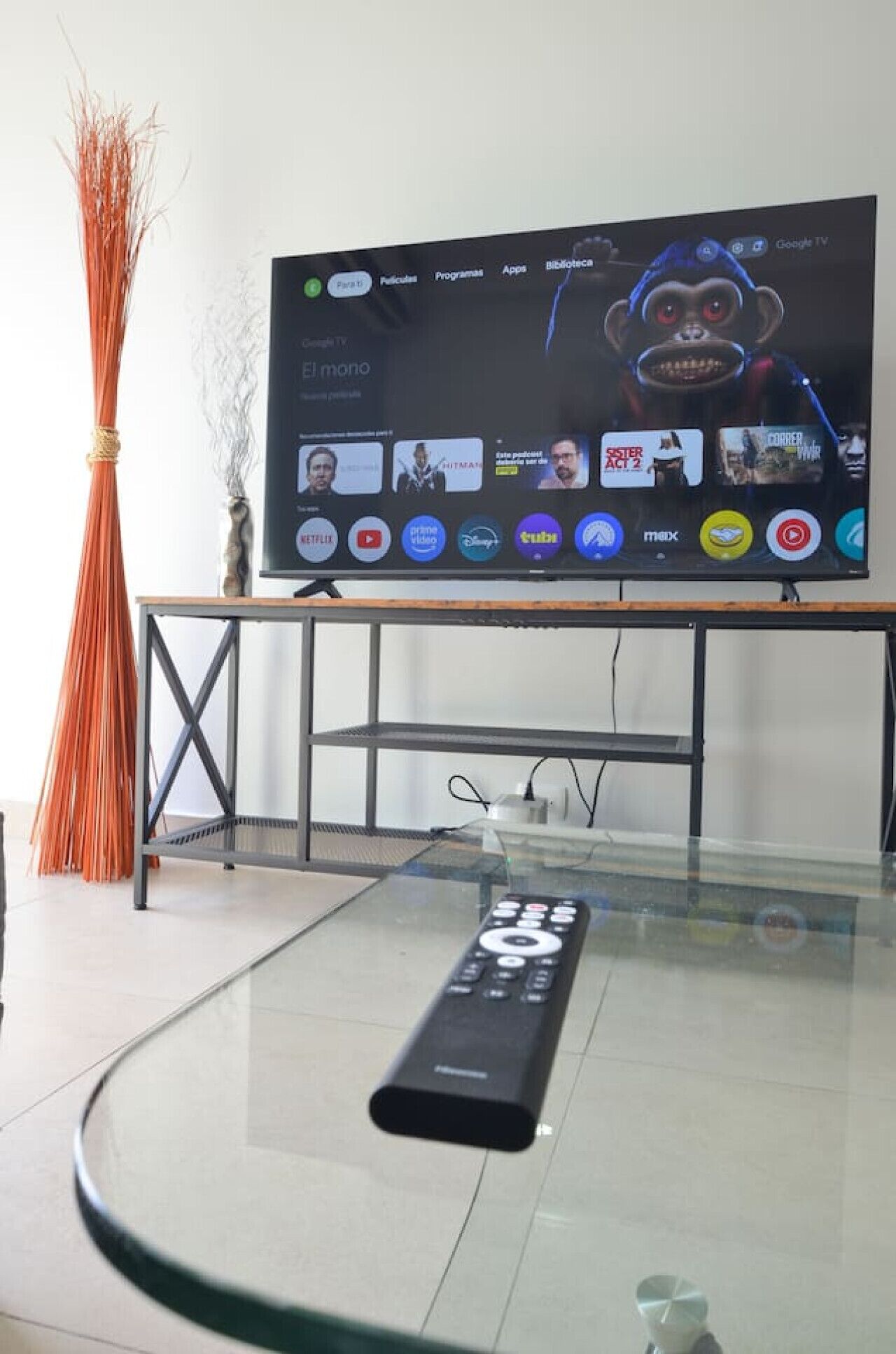 A modern living room with a glass coffee table, a remote control, and a flat-screen TV mounted on a wall.