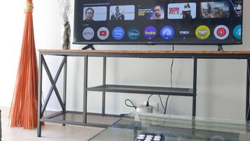 A modern living room with a glass coffee table, a remote control, and a flat-screen TV mounted on a wall.