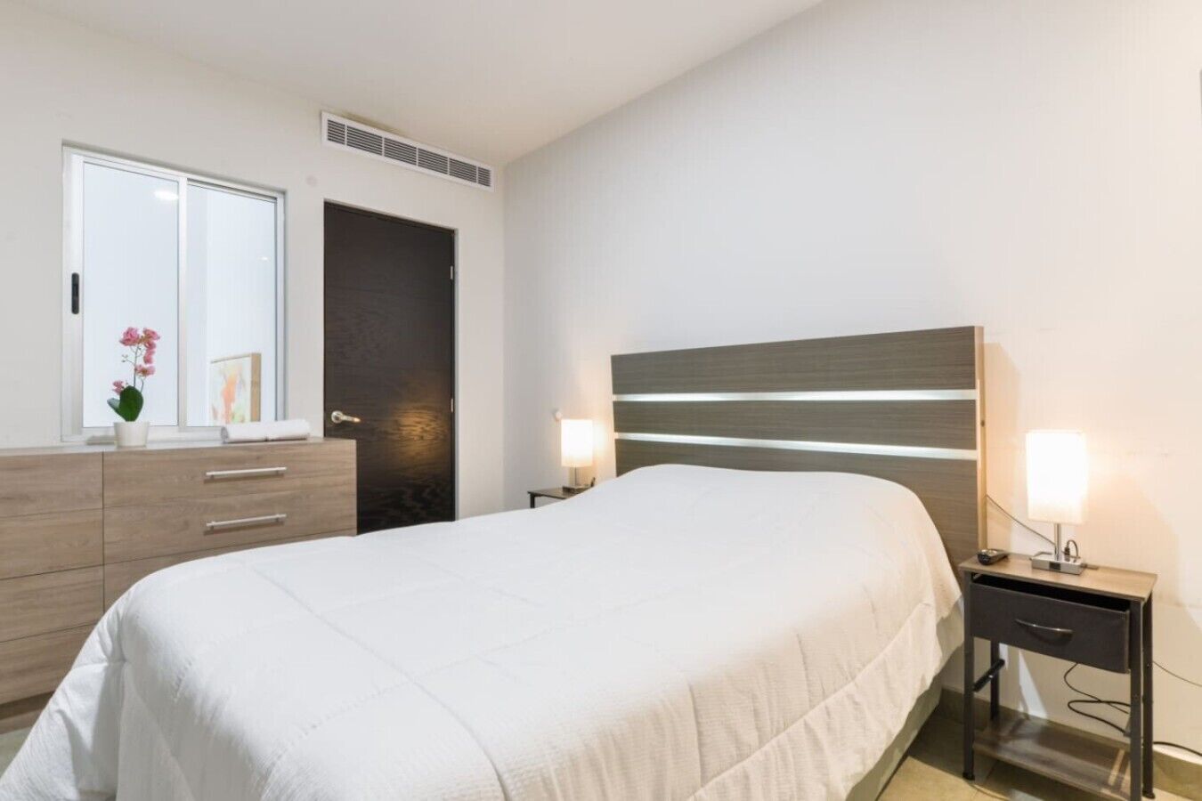Een slaapkamer met een bed, een dressoir, een spiegel, een deur en nachtlampjes.