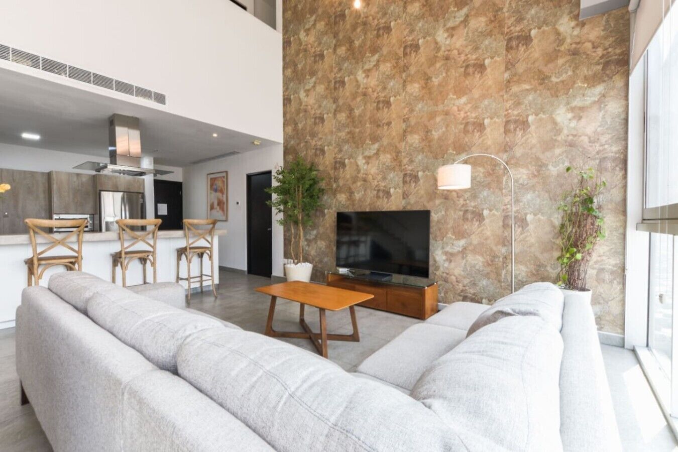 A modern living room with a stone accent wall, a flat-screen TV, a sofa, a dining area with bar stools, and a kitchen in the background.