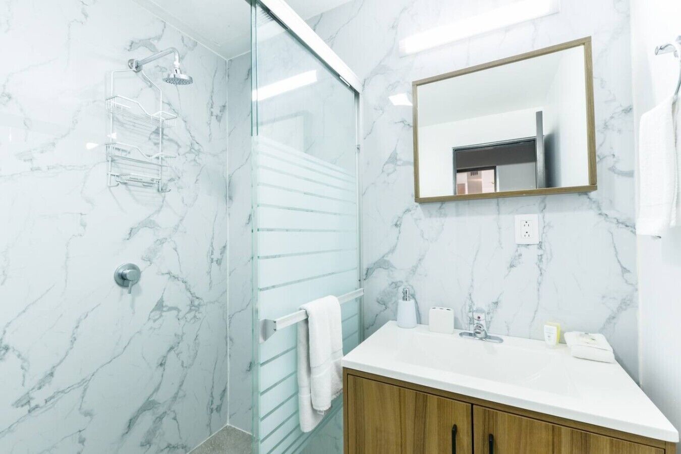 A bathroom with a glass shower enclosure, a white marble wall, a wooden vanity, and a mirror.