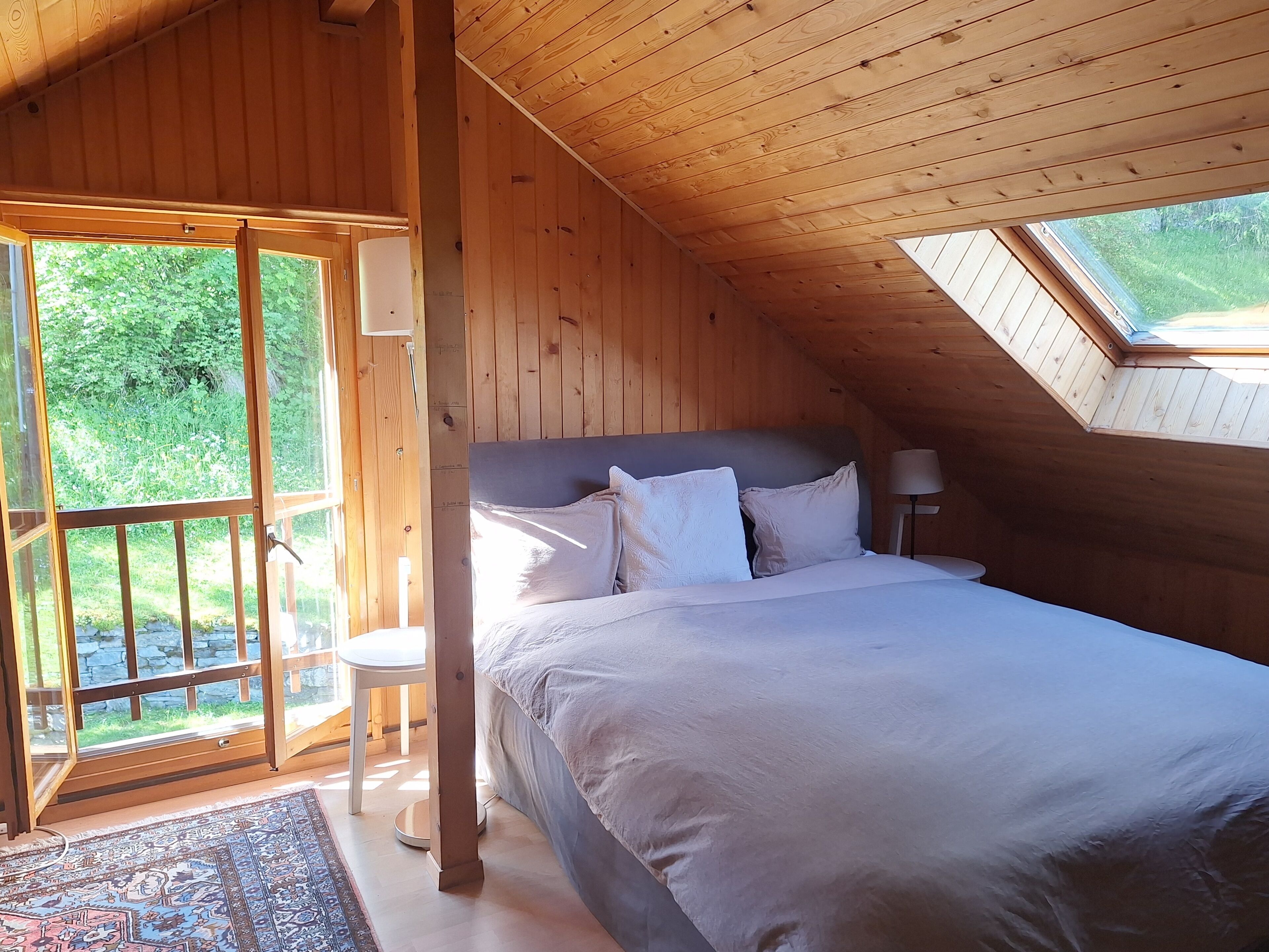Un dormitorio de cabaña de madera con cama, mesita de noche, lámpara y vistas a la vegetación a través de un ventanal grande.
