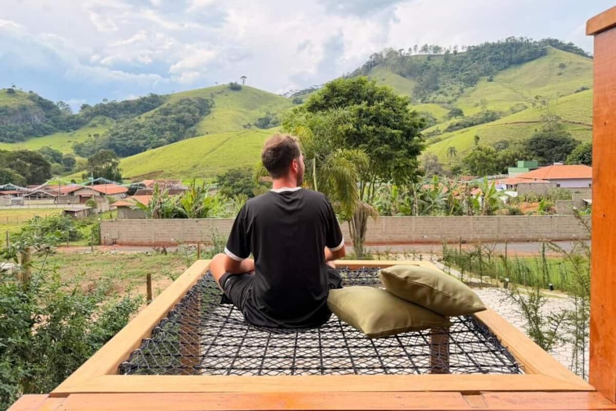 Uma pessoa sentada em uma rede, com vista para colinas verdes e uma aldeia ao fundo.