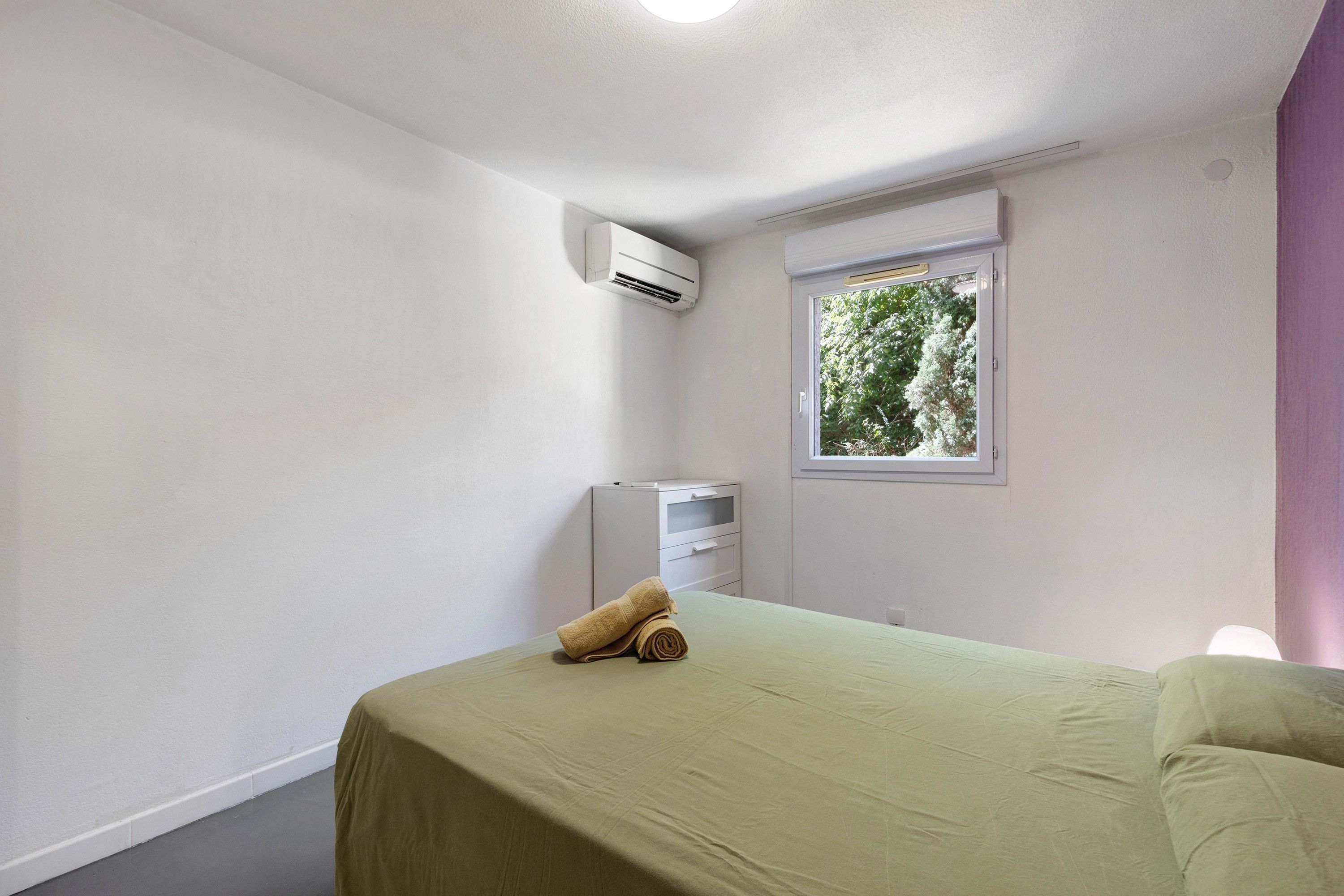 A room with a bed, an air conditioning unit, a window with a view of trees, and a small cabinet.