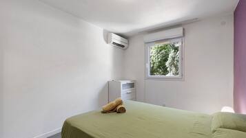 A room with a bed, an air conditioning unit, a window with a view of trees, and a small cabinet.