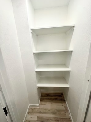 En smal hall med vita väggar och trägolv leder till en vit garderob med tre hyllplan.
