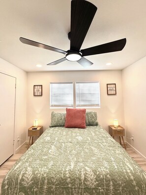 Ein Schlafzimmer mit einem Bett, zwei Nachttischlampen, einem Deckenventilator und zwei Fenstern mit Jalousien.
