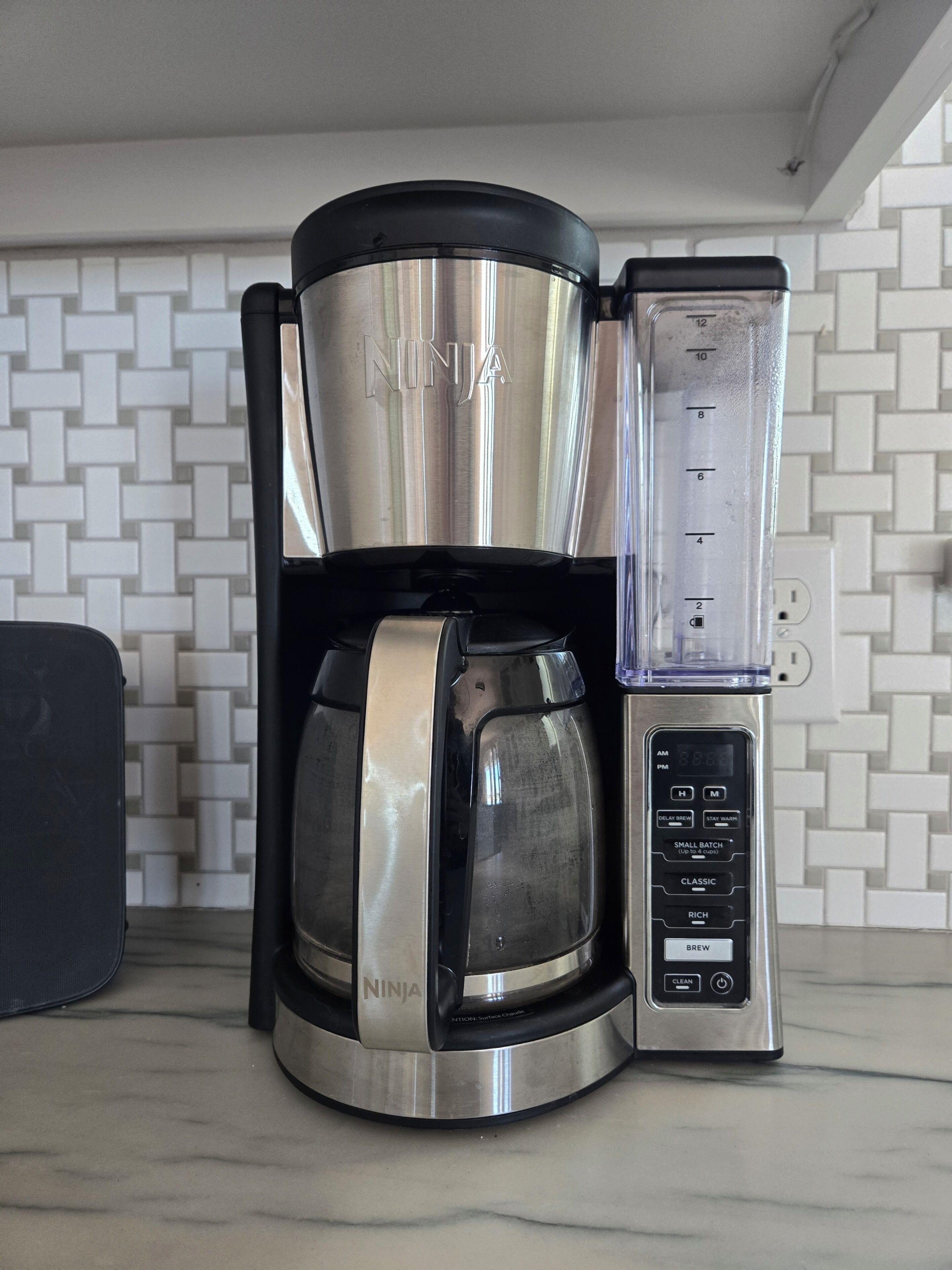 A NINJA coffee maker with a digital display, a carafe, and a drip tray, set on a kitchen counter with a white tiled backsplash.