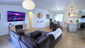 A modern living room with a grey sofa, a flat-screen TV, a dining area with a table and chairs, and a kitchen area with white cabinets and a microwave.