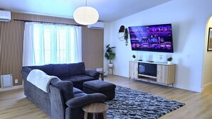 A living room with a dark sofa, a flat-screen TV, a wooden cabinet, and a large window with curtains.