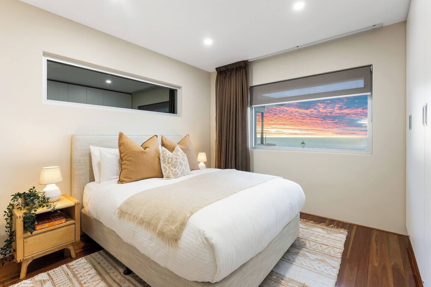 Une chambre à coucher avec un lit, des tables de chevet, une petite table de nuit avec une lampe, une fenêtre donnant sur un coucher de soleil et un luminaire au plafond.