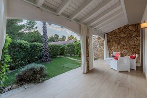 Eine überdachte Terrasse mit weißen Korbmöbeln, einer Steinwand und Blick auf einen gepflegten Garten.