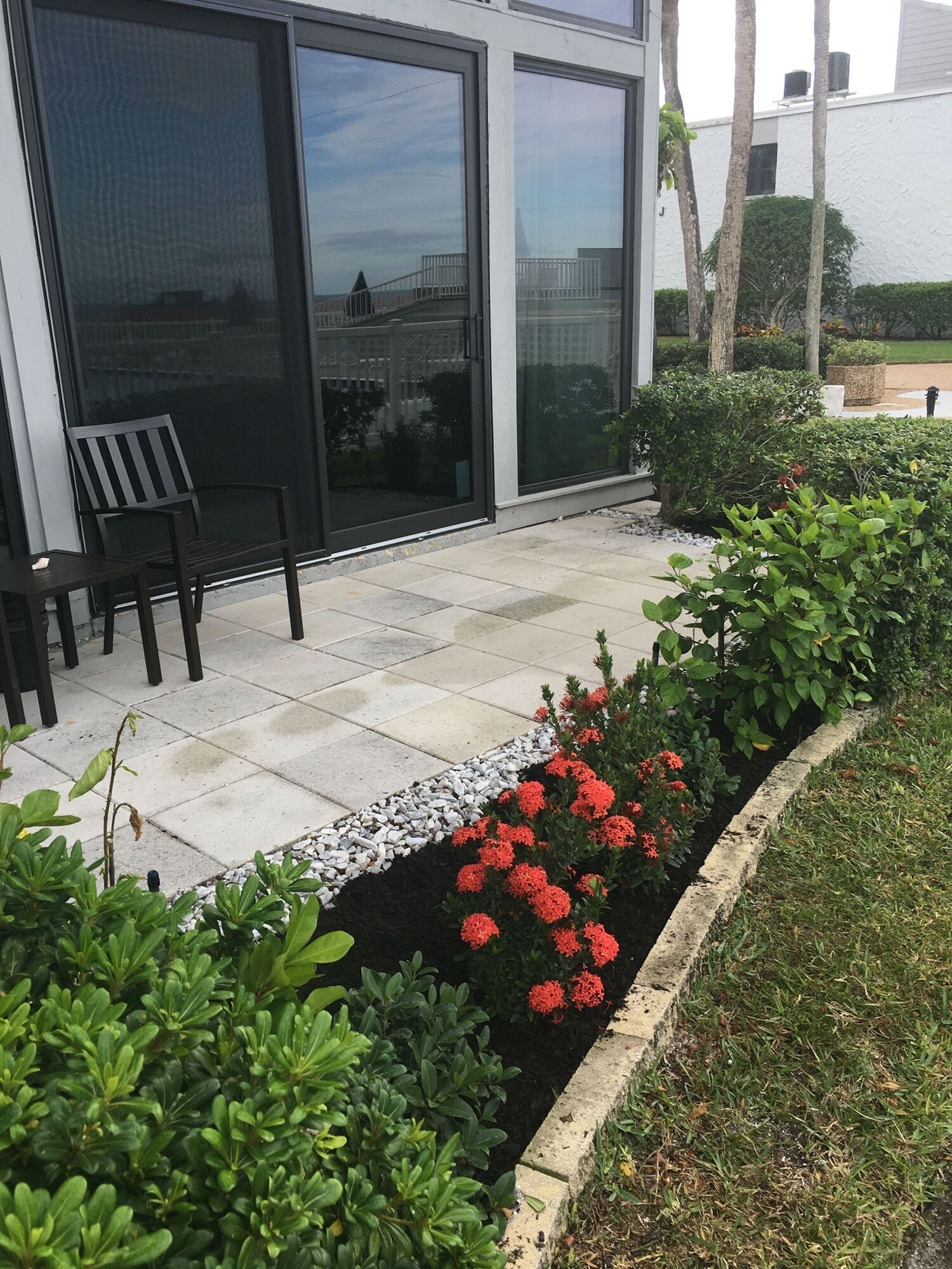 A modern building with large glass doors, a patio with a bench, and a garden with red flowers.
