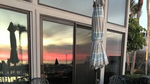 Une terrasse extérieure avec une table et des chaises, un parasol à rayures et une vue sur le coucher du soleil à travers de grandes fenêtres.