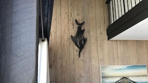 Un salon chaleureux avec un plafond en bois, une tête d’animal fixée au mur, une lampe, un canapé et un tableau encadré représentant une jetée.